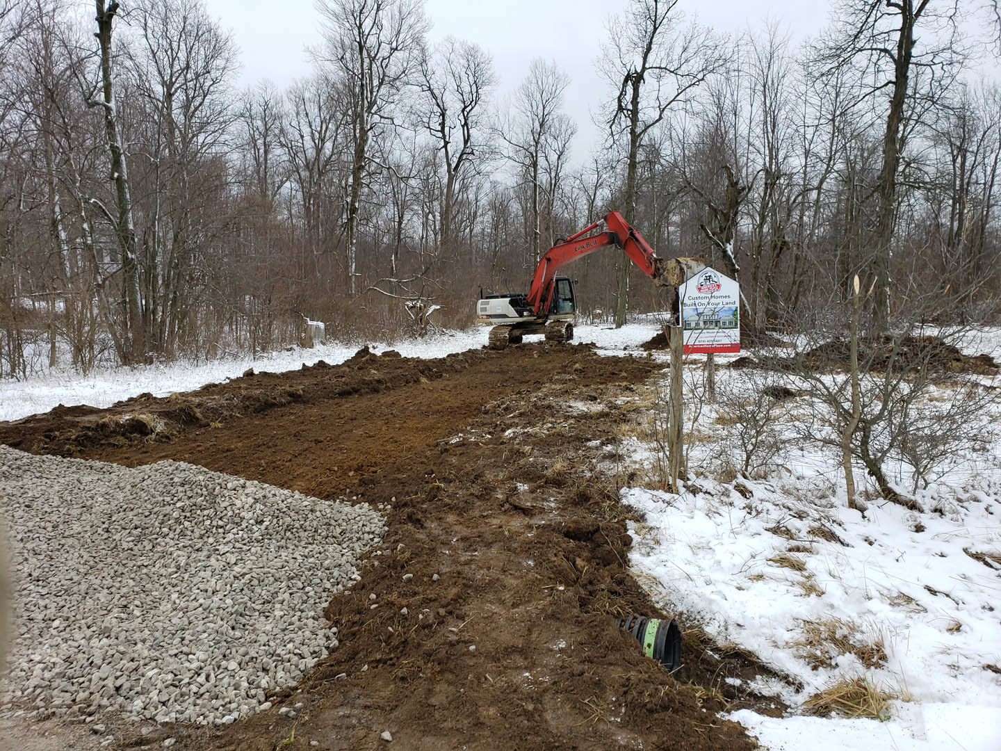 Snow-covered construction site with red excavator, crane, pile of white rocks, dirt road, and sign displaying a house, surrounded by winter trees.