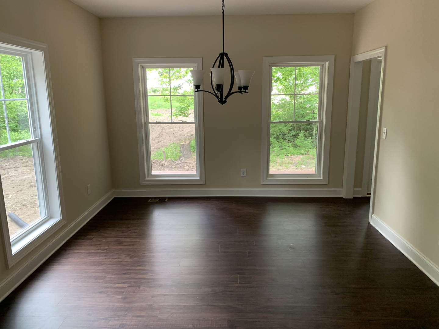 Chandelier hanging from ceiling above dark hardwood floor, large windows with views of green trees, neutral walls, natural daylight filling the room