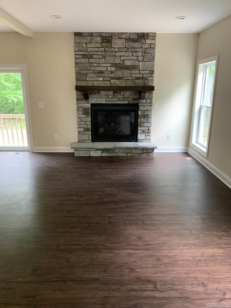 Stone fireplace with wood mantel set against a white wall, wood flooring, large window with wooden railing overlooking trees