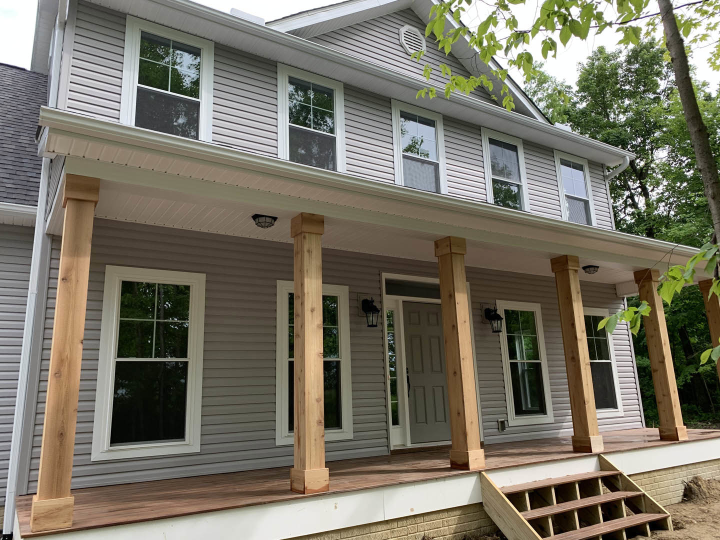 Two porches with white railings, white-framed windows reflecting trees, white door with black handle, light siding, and wooden staircase leading to entry.