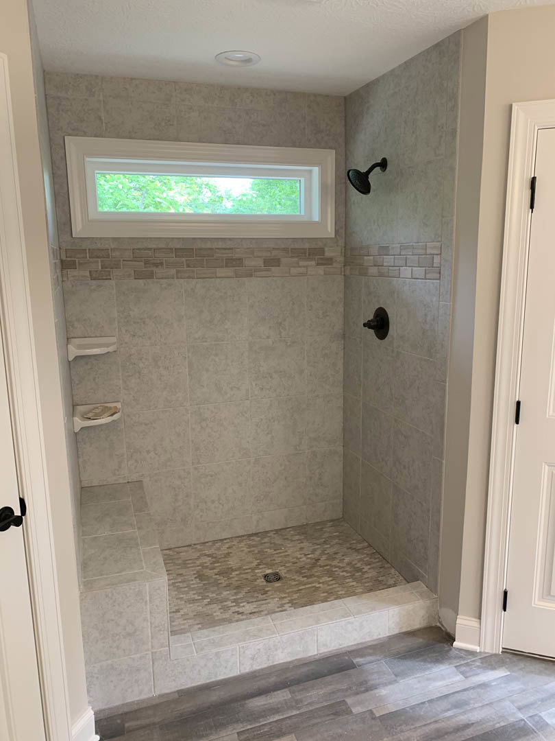 Shower with a white-framed window, wall-mounted shower head, wood flooring, and shower drain in a bathroom with plaster walls