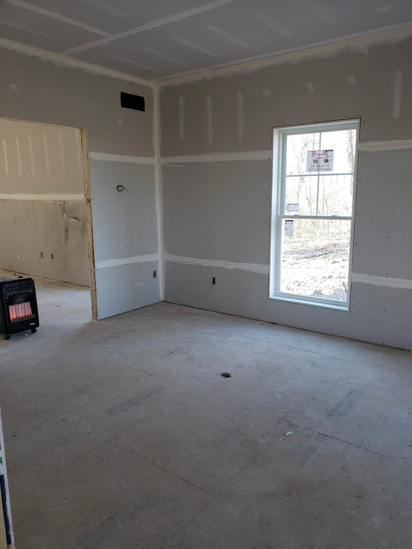 White-painted room with a large window, black rectangular stove featuring visible red flame, light flooring, and a wall-mounted sign with logo.
