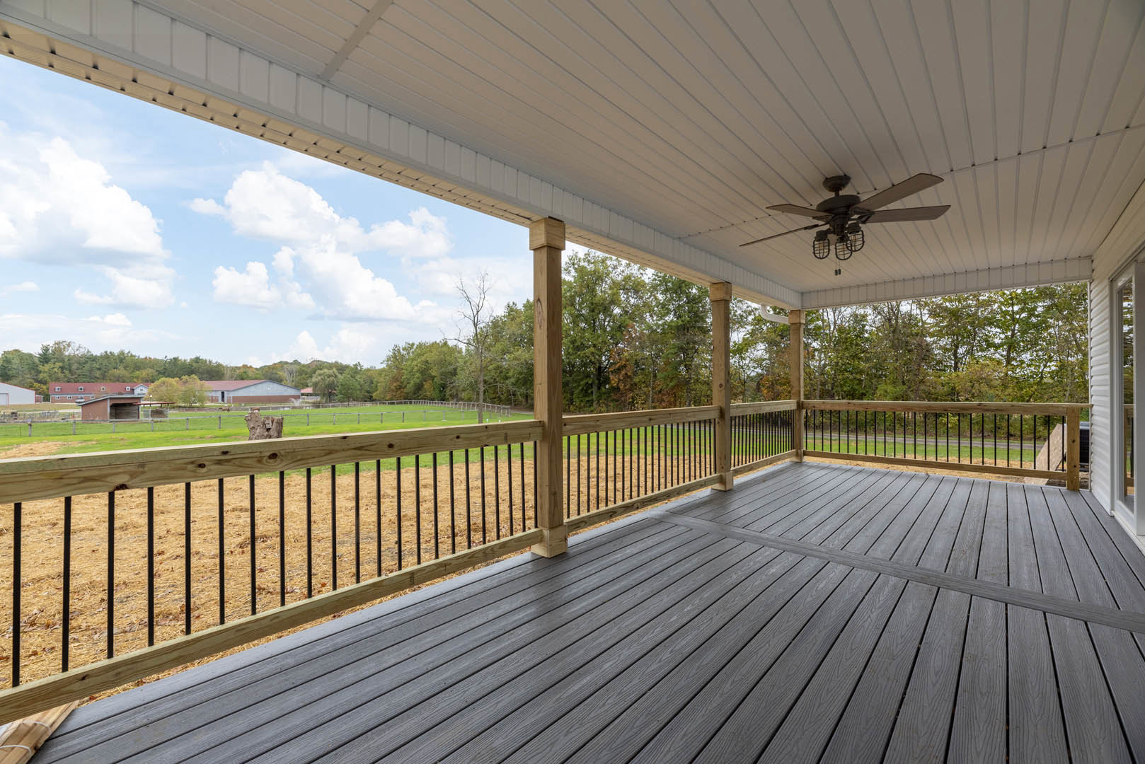 Wooden deck with white ceiling, ceiling fan, wooden pillars, railing, and expansive grass field bordered by trees.