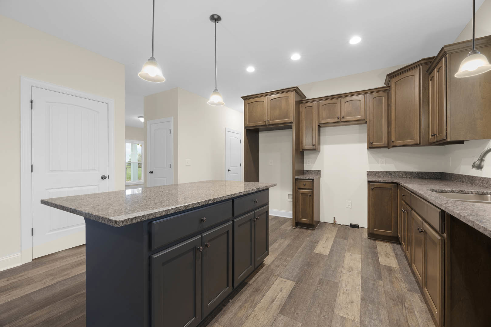 Spacious kitchen featuring a large central island with granite countertops, wooden cabinetry, tile flooring, stainless steel sink, and white door in the background.