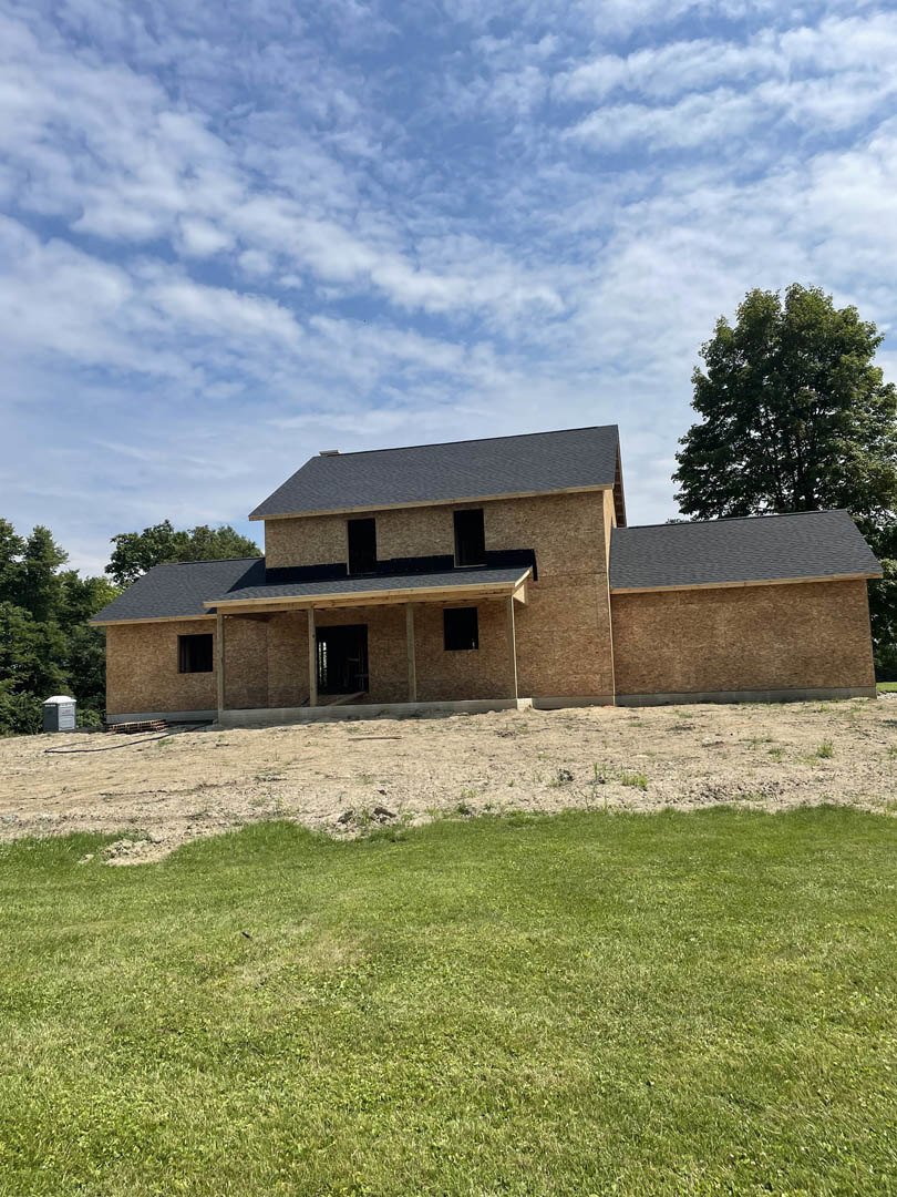 Modern two-story house with light siding, large windows, and a manicured grassy lawn bordered by mature trees under a bright blue sky