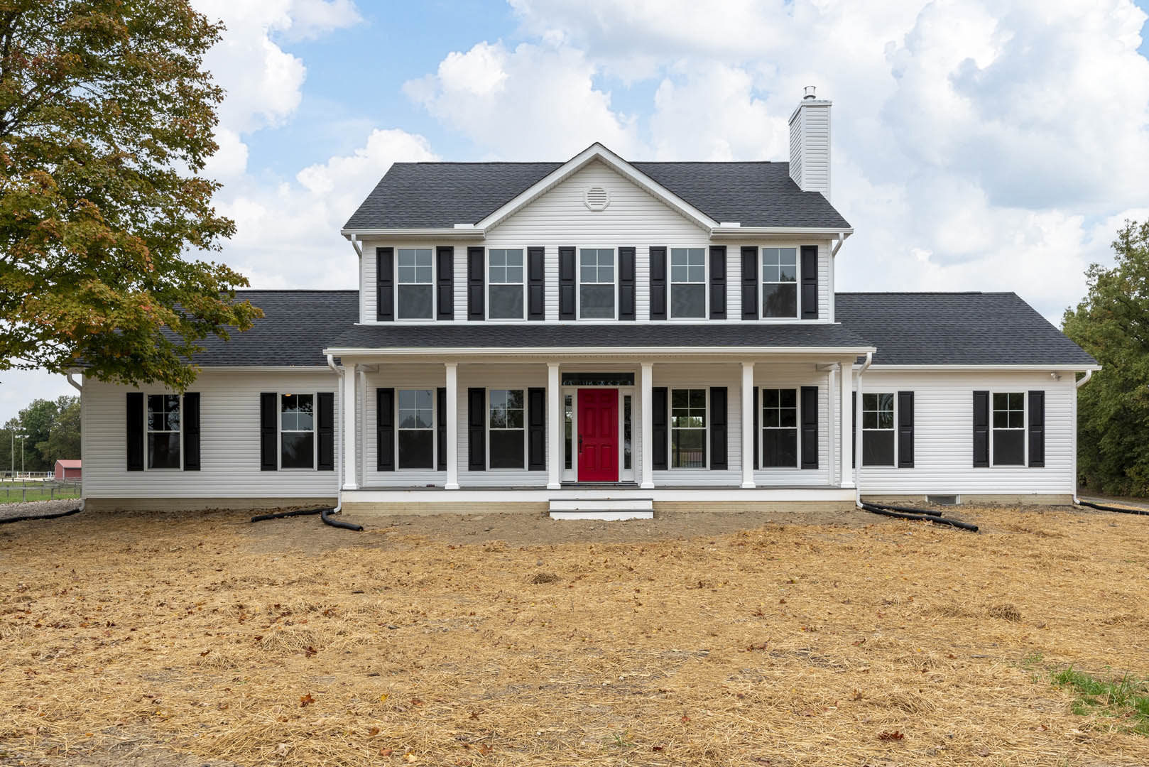 White two-story house with red front door, white-framed windows, wide porch, brown grass lawn, and leafy green tree in foreground