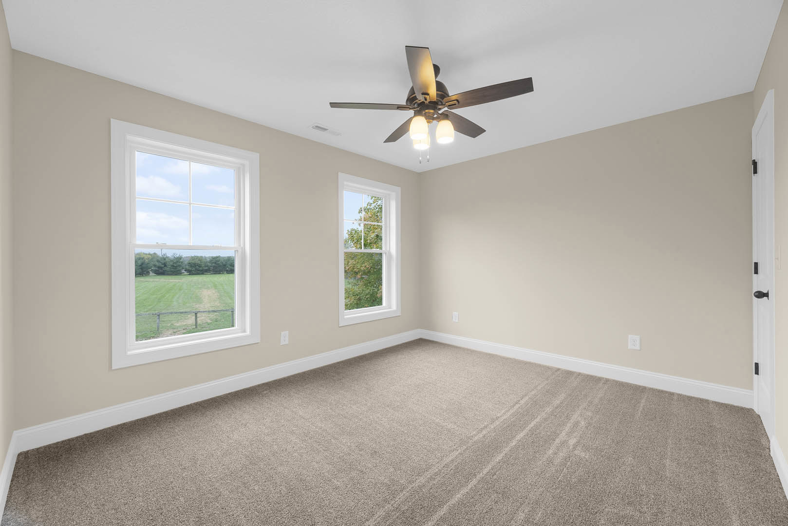 Carpeted room with white walls, ceiling fan with lights, large windows overlooking green lawn and trees