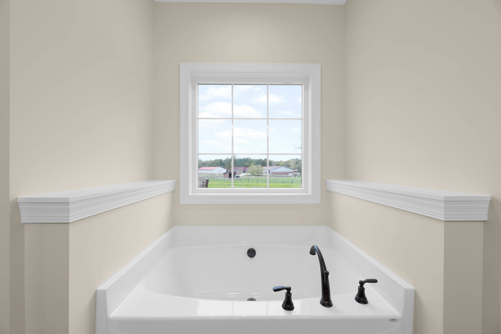 White freestanding bathtub with black faucet beneath large window overlooking farmland, white wall molding visible, decorative statue on windowsill