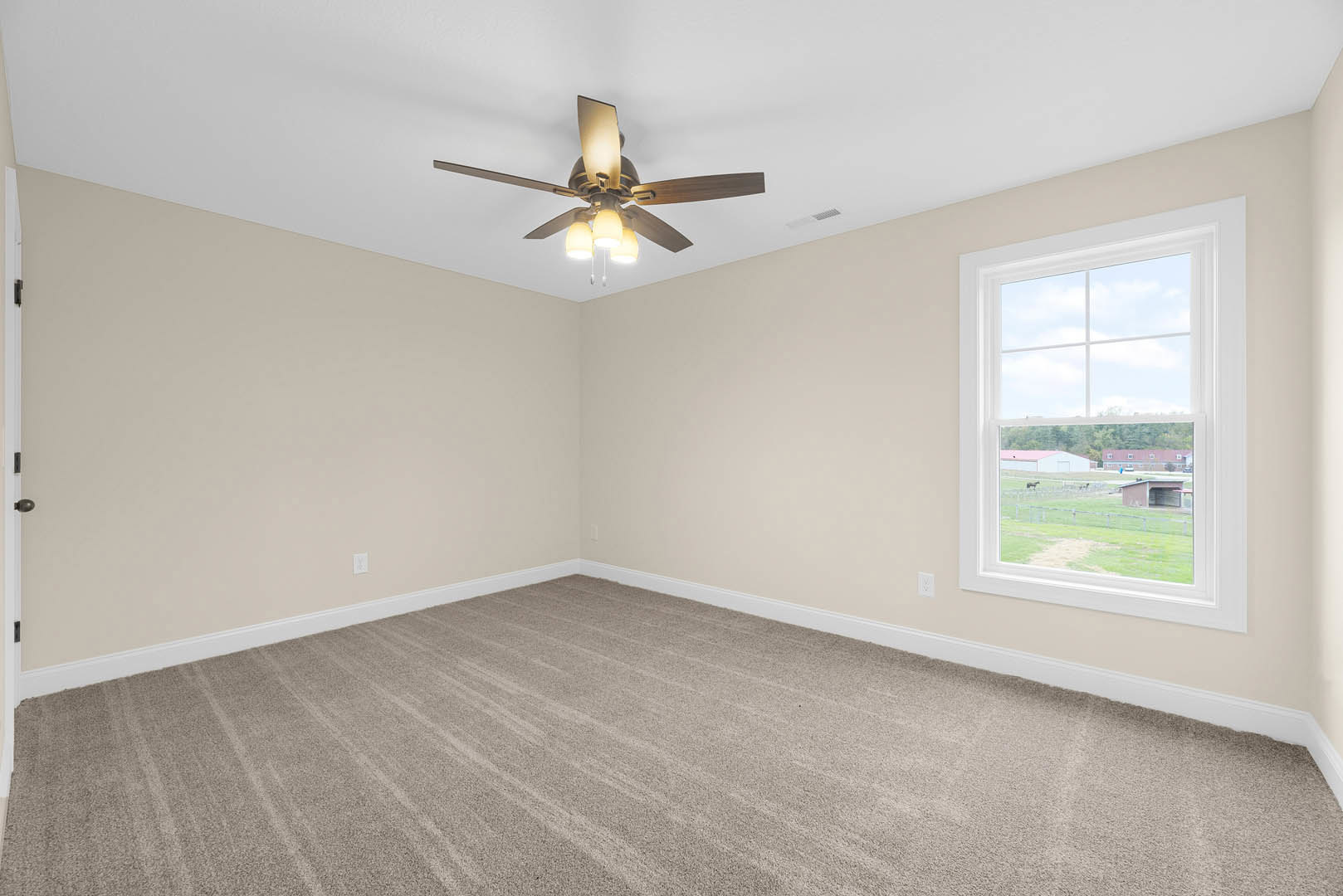 Carpeted room with white walls, ceiling fan with light fixture, large window overlooking farmland