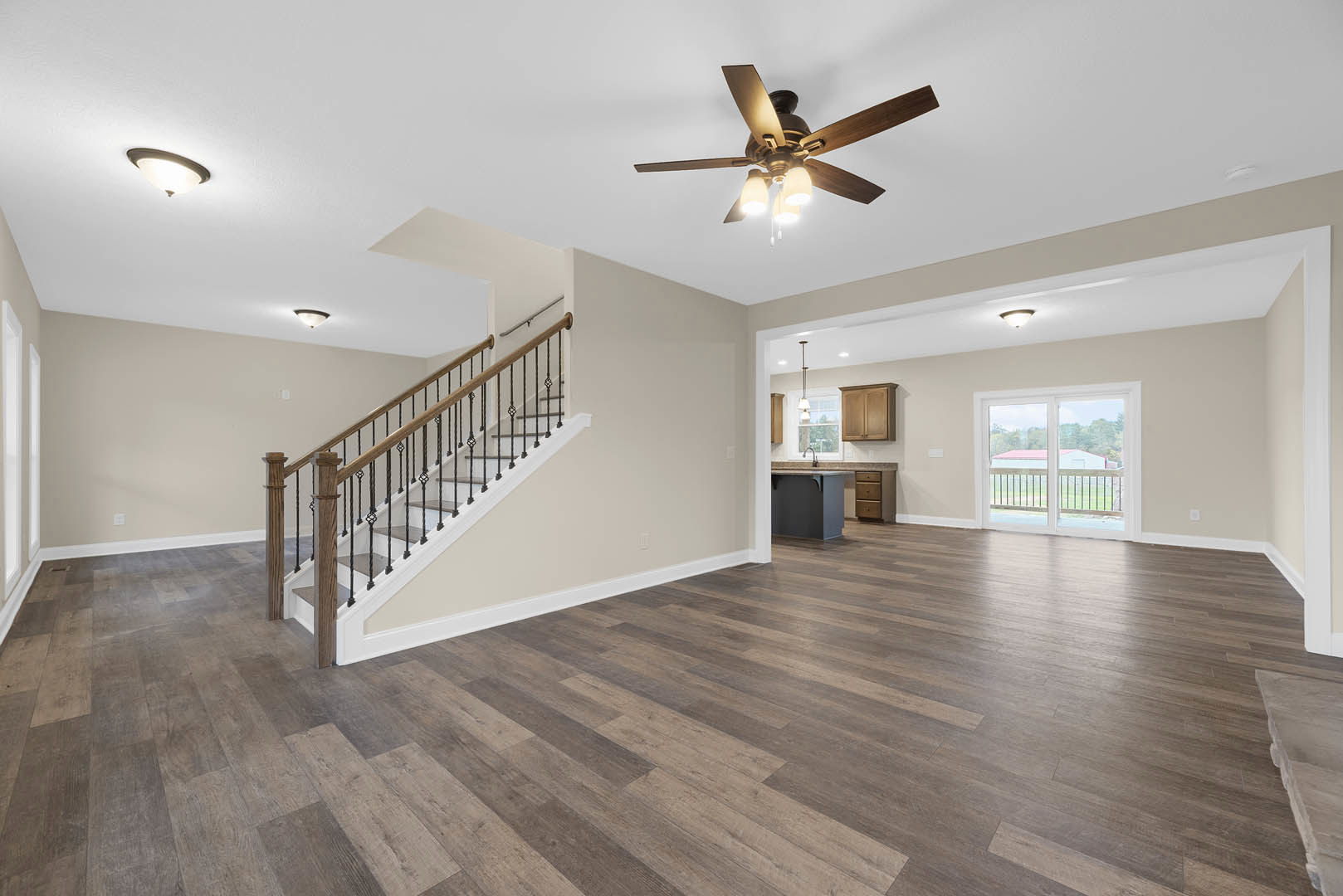 Open living area with wood flooring, metal-railed staircase, ceiling fan with lights, brown cabinet against white wall, and window with railing