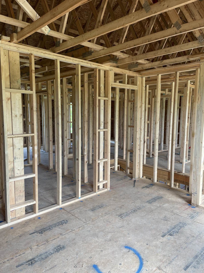 Exposed wooden beams and framing inside a partially constructed house, with construction materials and tire tracks visible on the subfloor.