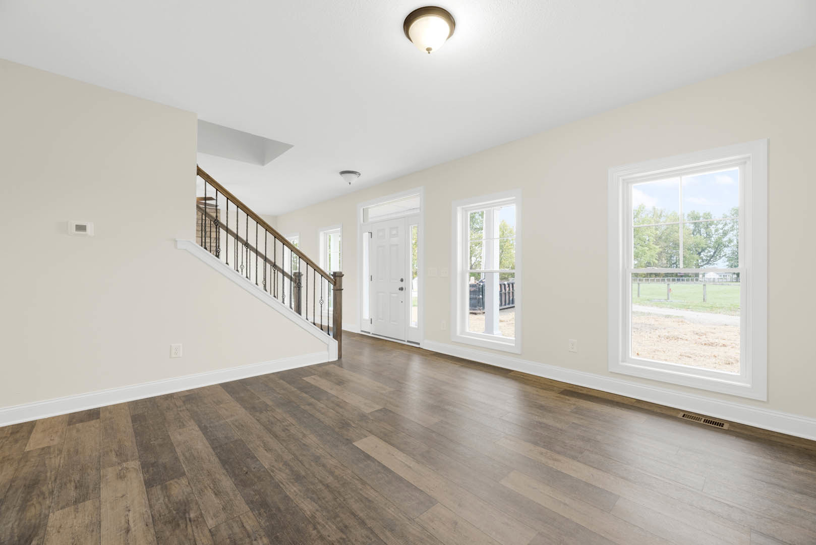 Open-concept room featuring hardwood floors, white walls with crown molding, a wooden staircase with black railings, large windows overlooking a field and trees, and a modern light