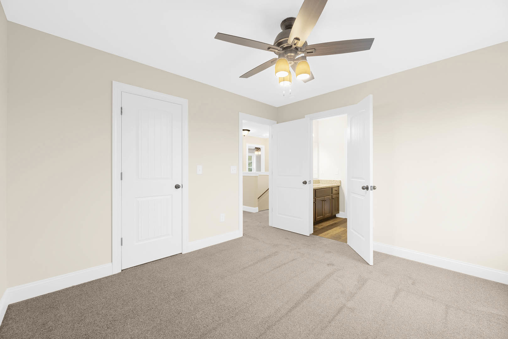 Carpeted room with white walls, ceiling fan with lights, two white doors with black handles, partial view of kitchen counter