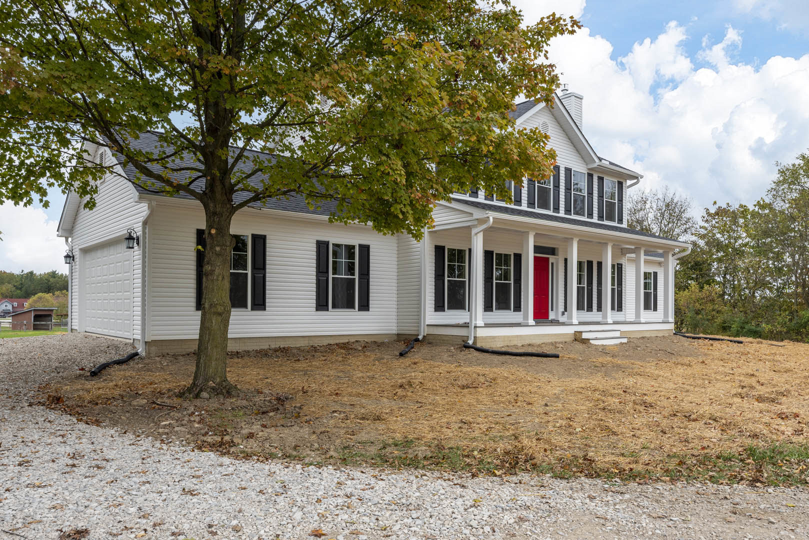White siding house with red front door, black shuttered window, white-framed windows, tree in yard, dirt patch and black pipe on ground