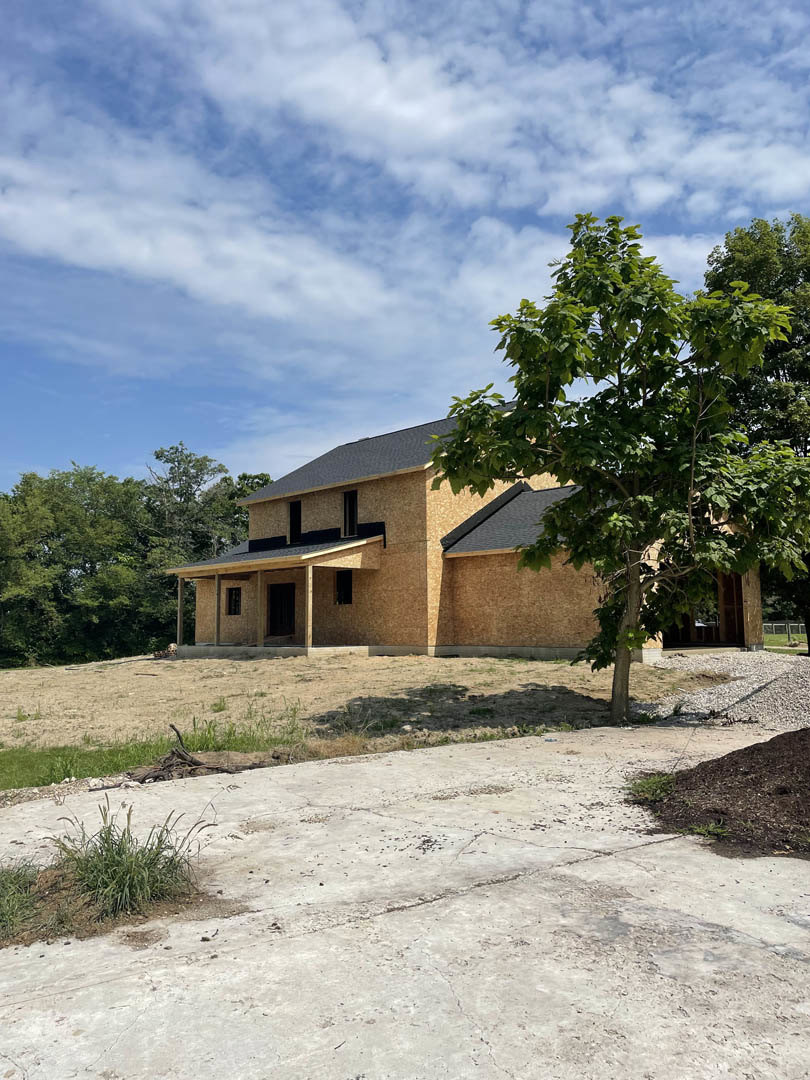 Modern house with light-colored siding, large windows, and a mature tree in the backyard; concrete walkway bordered by grass and landscaping under a partly cloudy sky