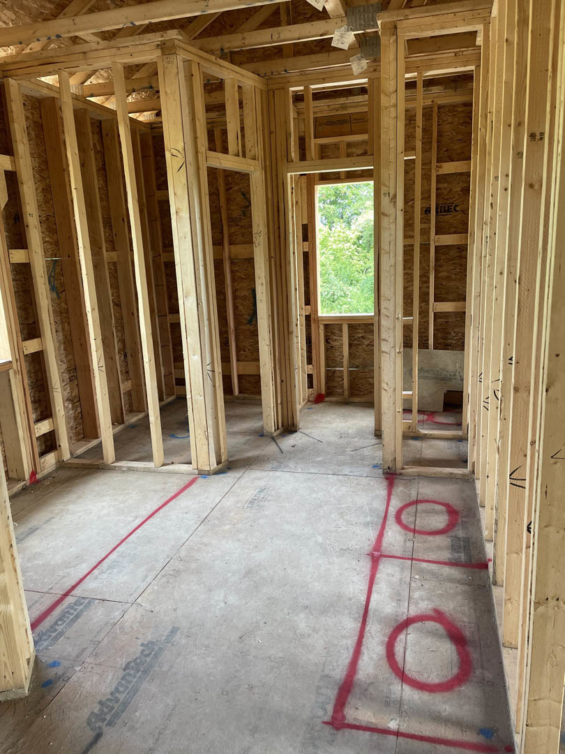 Room under construction featuring exposed wood ceiling beams, concrete floor marked with red tape lines and circles, large window overlooking trees in the background