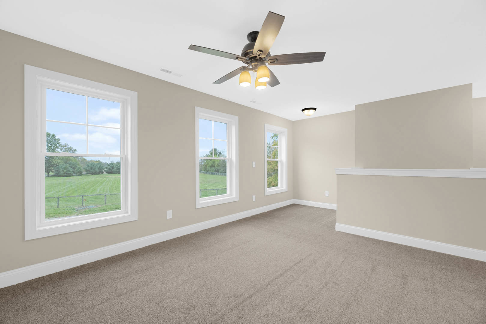Carpeted room with white walls, ceiling fan with two yellow lights, large windows overlooking a field and trees, crown molding along ceiling edges