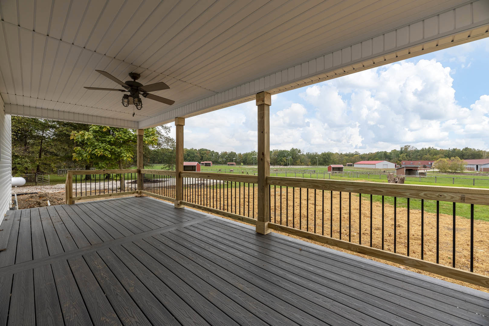 Wooden deck with railing and ceiling fan, covered by a roof, overlooking a fenced grassy yard with trees and clouds in the background.