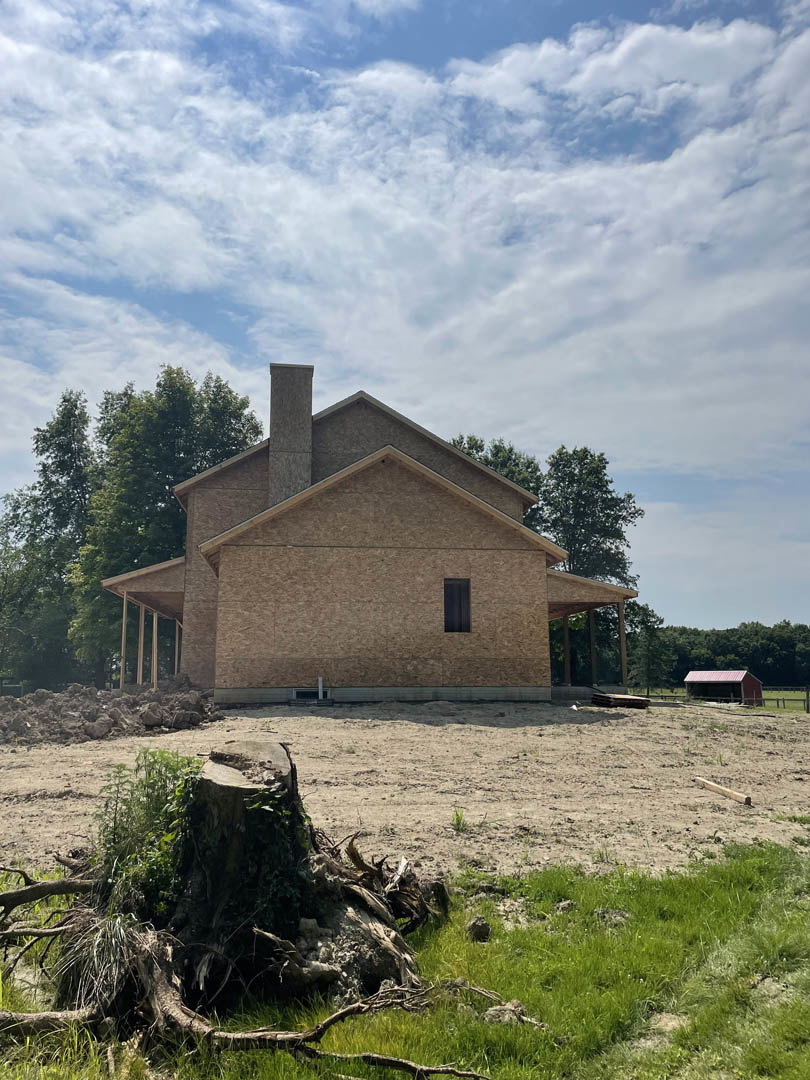 Framed custom home under construction with exposed wooden beams, unfinished walls, and surrounding trees in the background