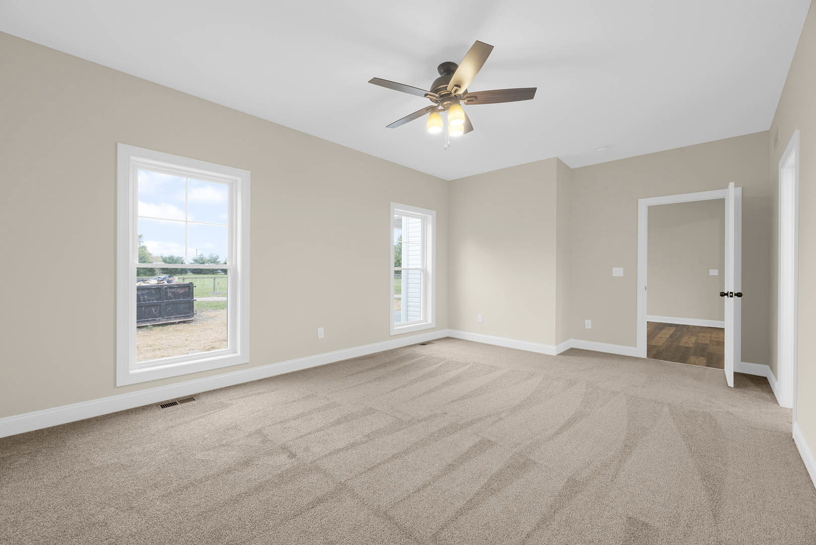 Carpeted room with white walls, ceiling fan with light fixture, two windows with white frames, white door with black handle, outdoor bin visible through window