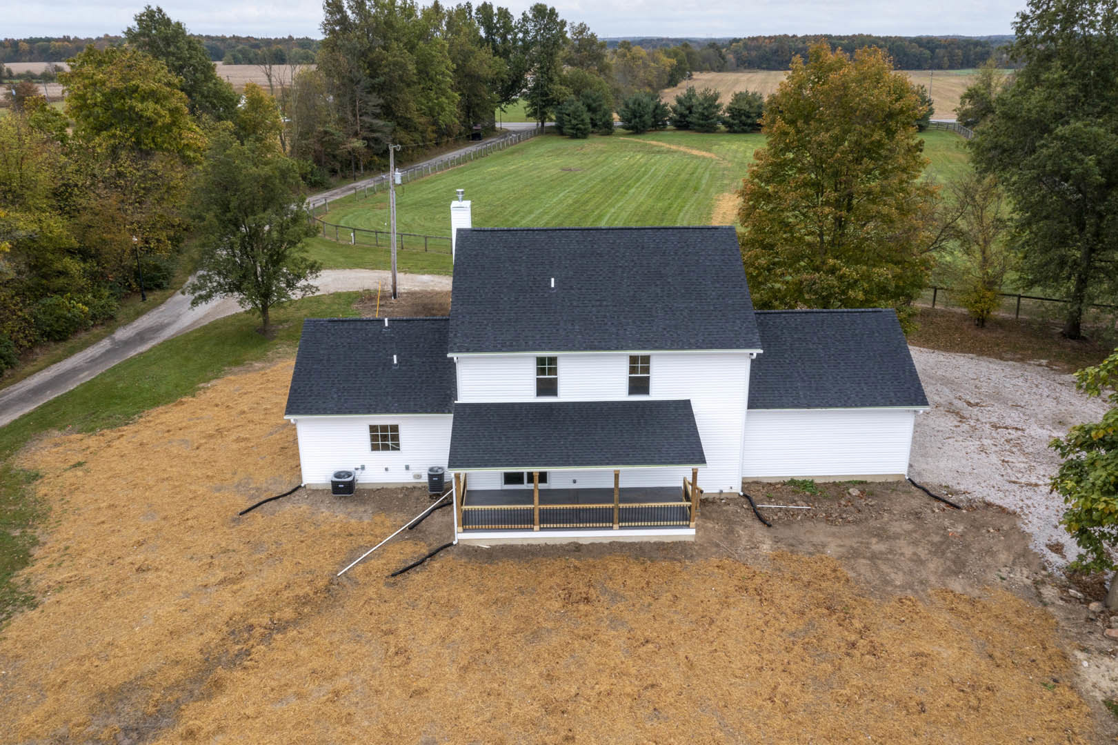 Two-story cottage with gray shingle roof, white porch railing, wooden fence, and mature leafy trees surrounded by green grass
