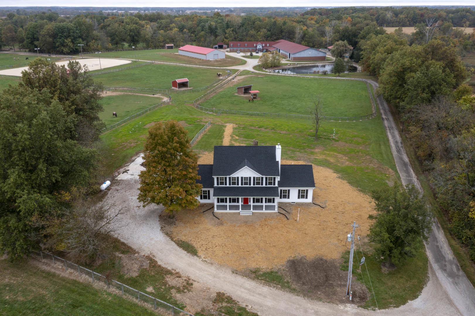 Modern house with black roof and light siding set in expansive grassy field, surrounded by mature leafy trees under open sky