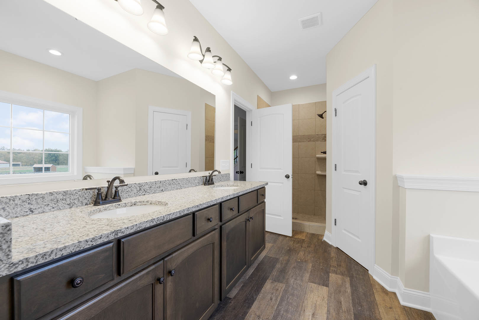 Bathroom with double vanity sinks, wood flooring, granite countertops, white cabinetry, silver faucet fixtures, and a large window providing natural light
