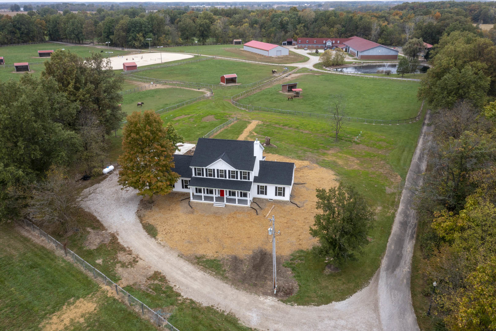 Two-story house with pink roof and red door, surrounded by grassy field, mature leafy tree, barn, and horse stable under open sky