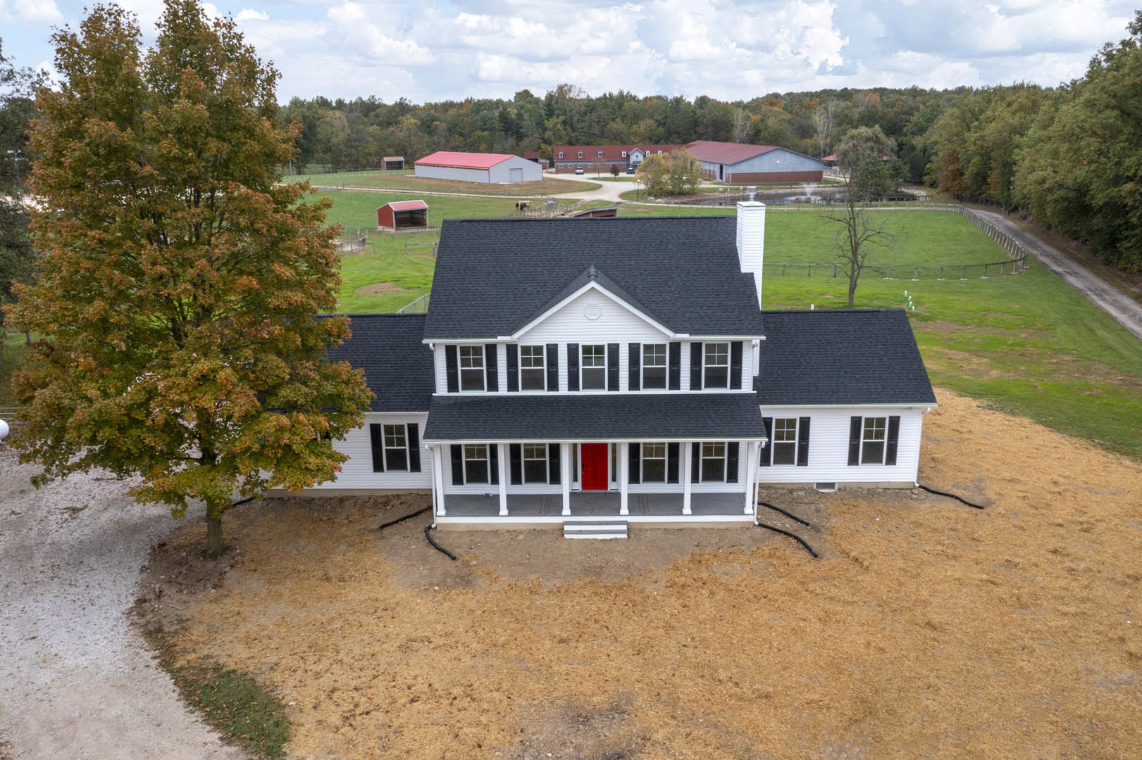 Two-story house with white siding, red front door, white trim, large grassy yard, mature leafy trees, cloudy sky overhead