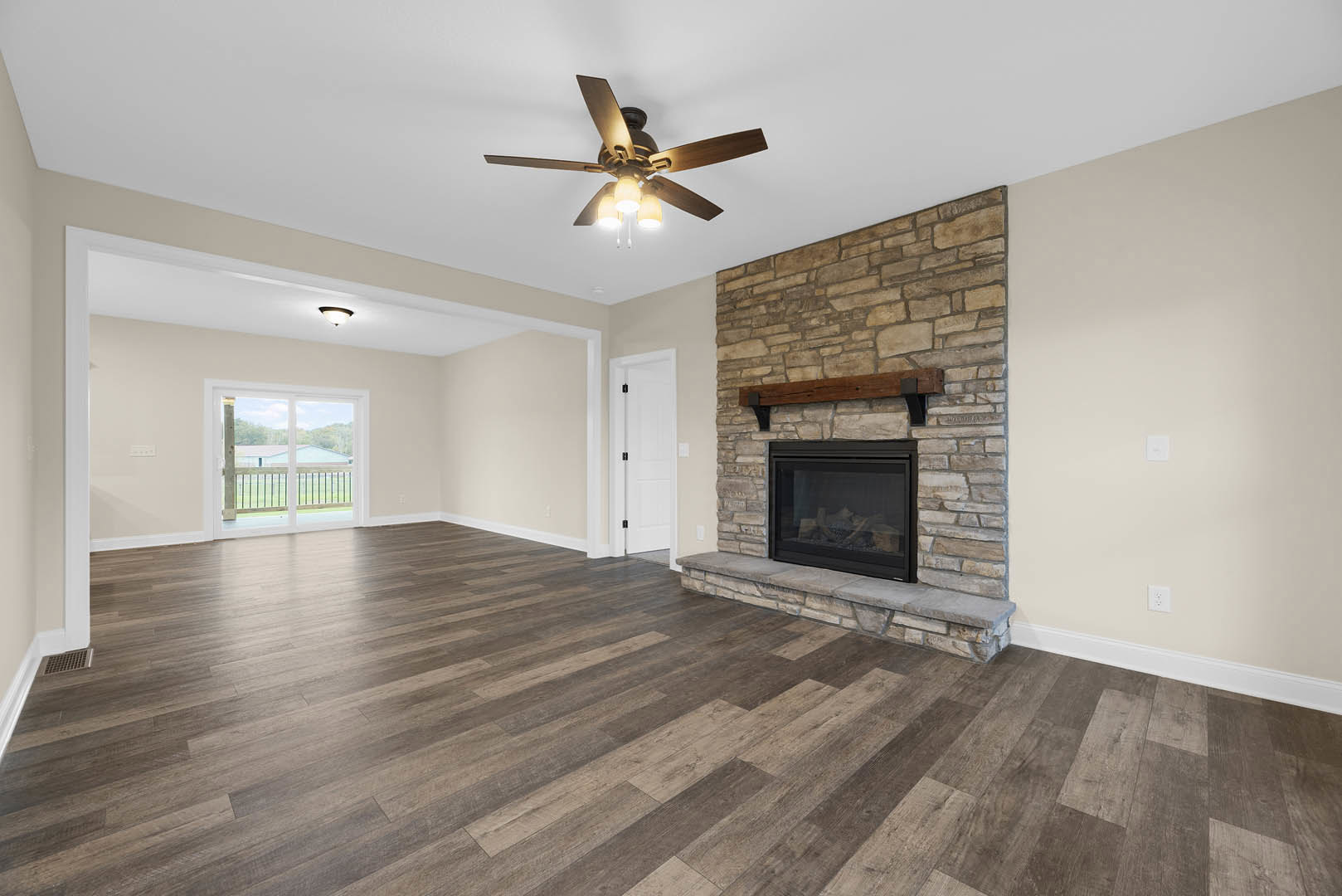 Living room with wood flooring, ceiling fan with lights, glass-door fireplace, white door with black handle, window overlooking railing and fence, plaster walls and ceiling