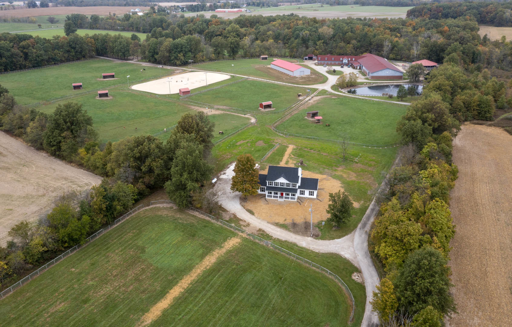 White house with red door and pink and white building set beside a large grassy field, bordered by trees and a fence, viewed from above