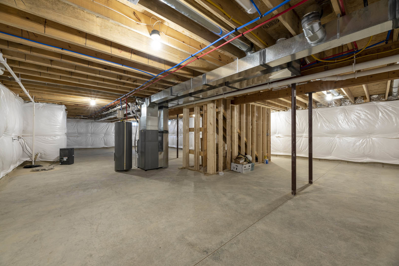 Exposed wood ceiling beams and metal pipes above a basement room, white plastic wall covering, black electrical box with wire, white sensor box with round top, blurry ceiling light