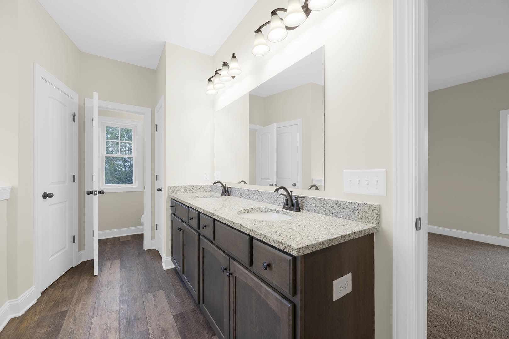 Bathroom with double sinks set in a white countertop, large mirror above, white cabinetry below, tile flooring, window with trees visible outside, white door with black knob