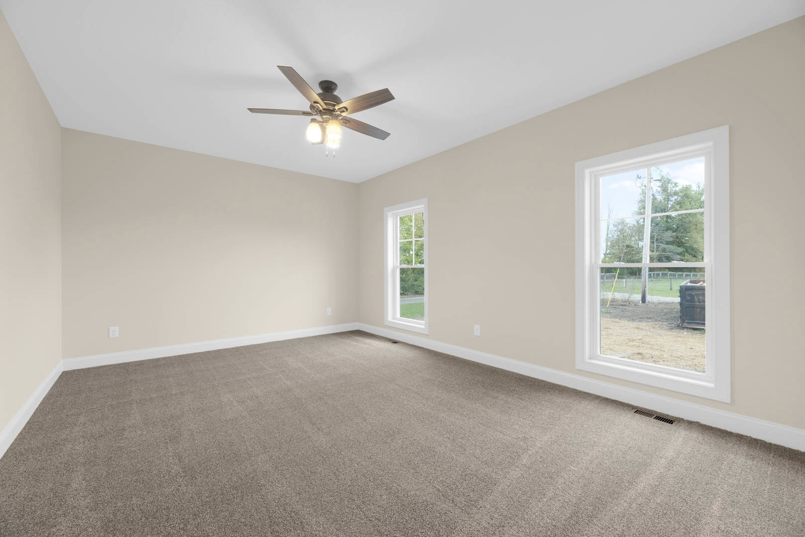 Carpeted room with white walls, ceiling fan with light fixture, large windows overlooking green yard and trees