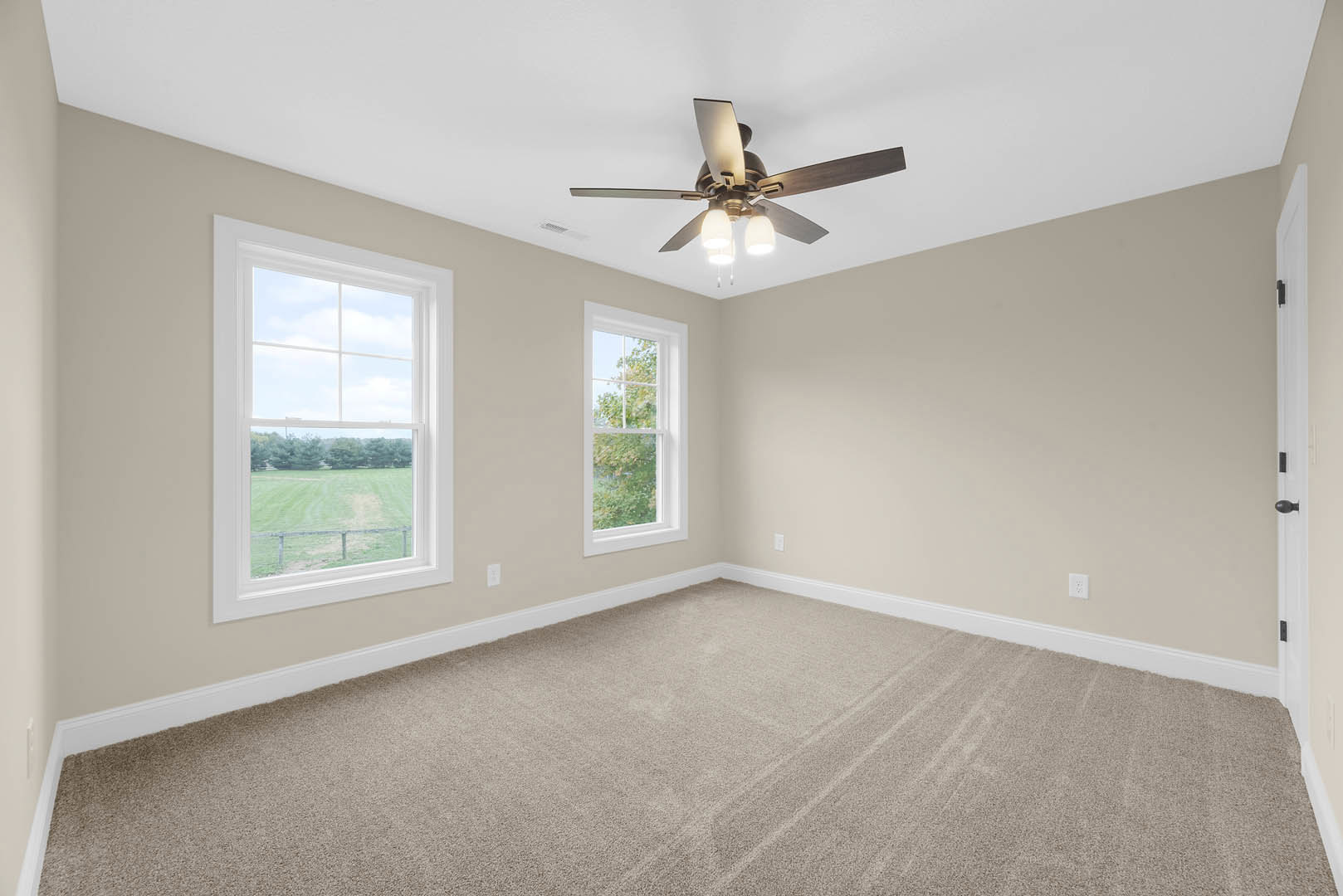 Carpeted room with white trim, ceiling fan with lights, two windows showing views of a grassy field and trees, plaster walls and ceiling