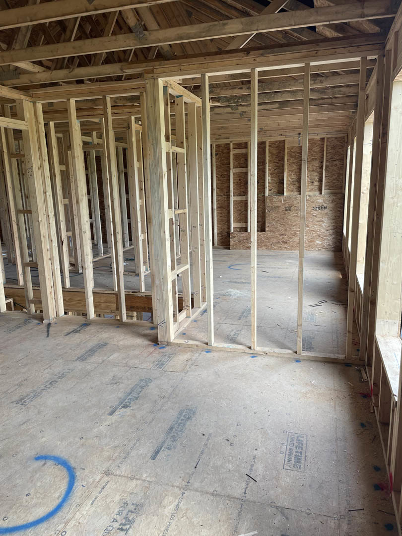 Exposed wood framing with beams and joists, unfinished floor, partial wall, blue marking on concrete, construction materials visible throughout room