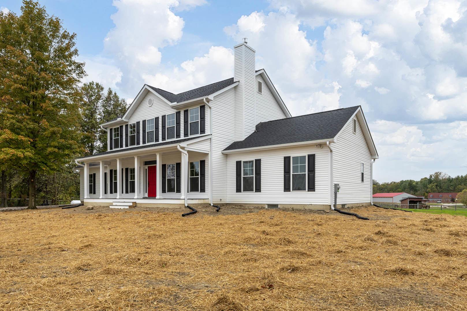 White siding house with black trim and shutters, red front door, leafy tree in front yard, brown grass field with black irrigation pipes, cloudy sky overhead