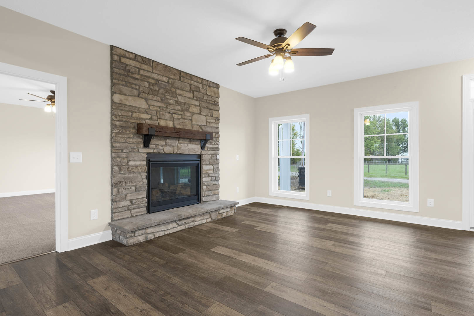 Stone fireplace with glass door, wood flooring, ceiling fan with lights, large window overlooking field and trees, plaster walls