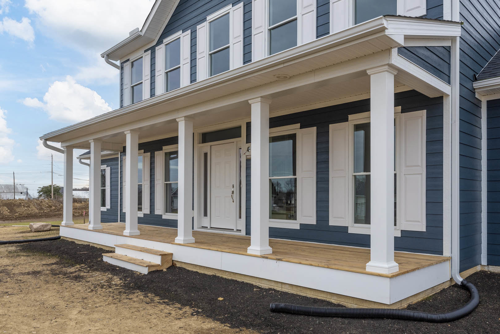 Two-story home with light-colored siding, spacious front porch, white door with silver handle, black pipe on ground, tree with power lines in background, concrete steps leading to