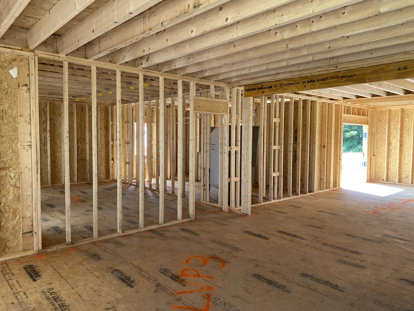 Exposed wooden beams and framing inside a partially constructed house, orange paint marking the concrete floor, unfinished ceiling and walls.