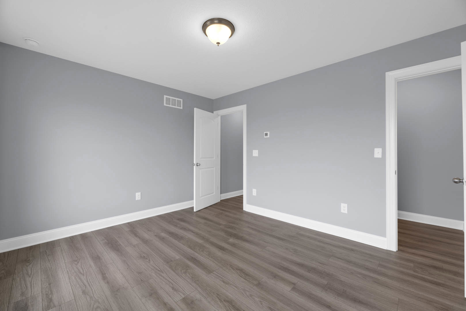 White paneled door with silver knobs open to a room featuring wood flooring, white baseboards, plaster walls, ceiling light fixture, and wall vent