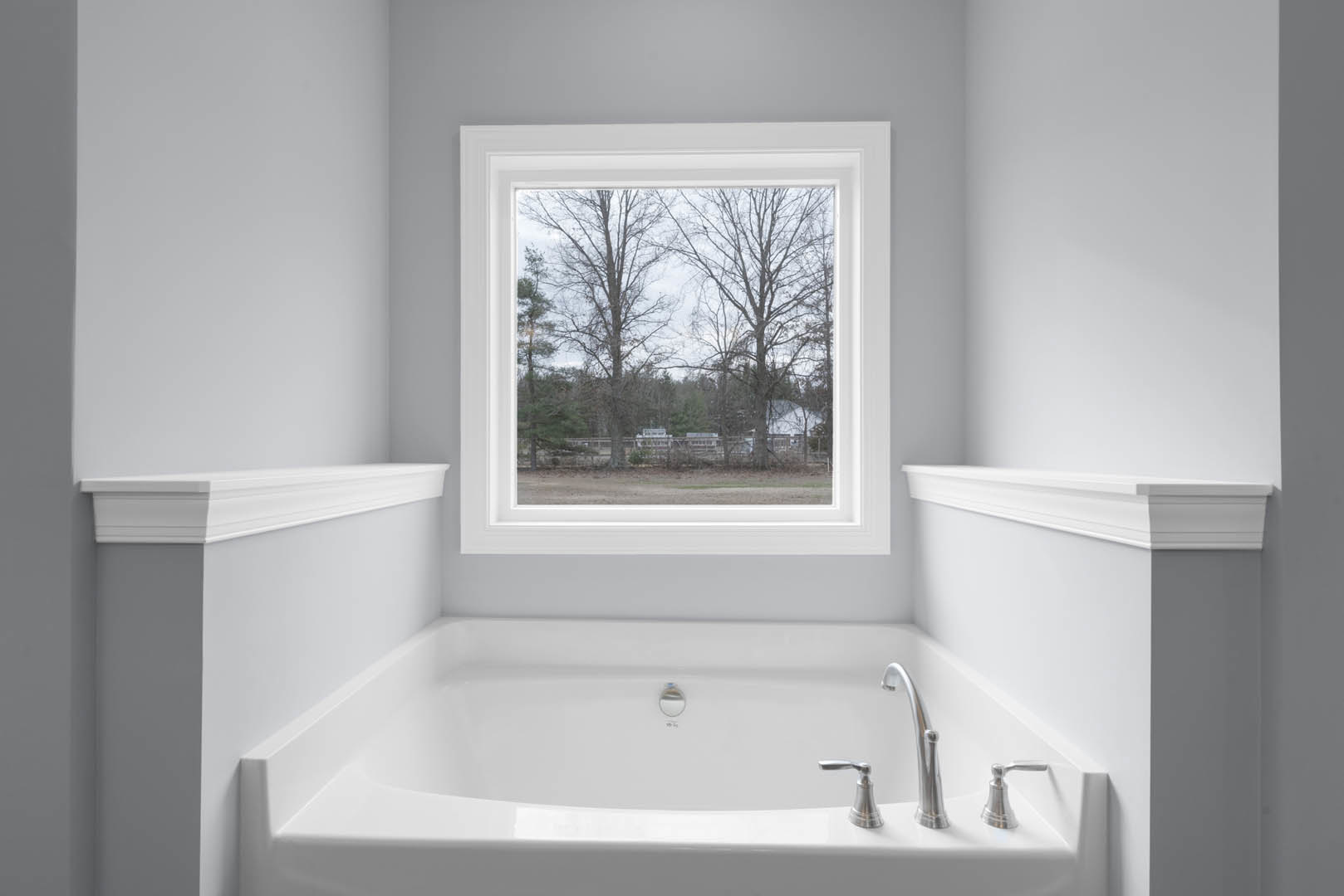 White freestanding bathtub beneath large window with tree views, chrome faucet and handle, smooth white wall, bright natural light