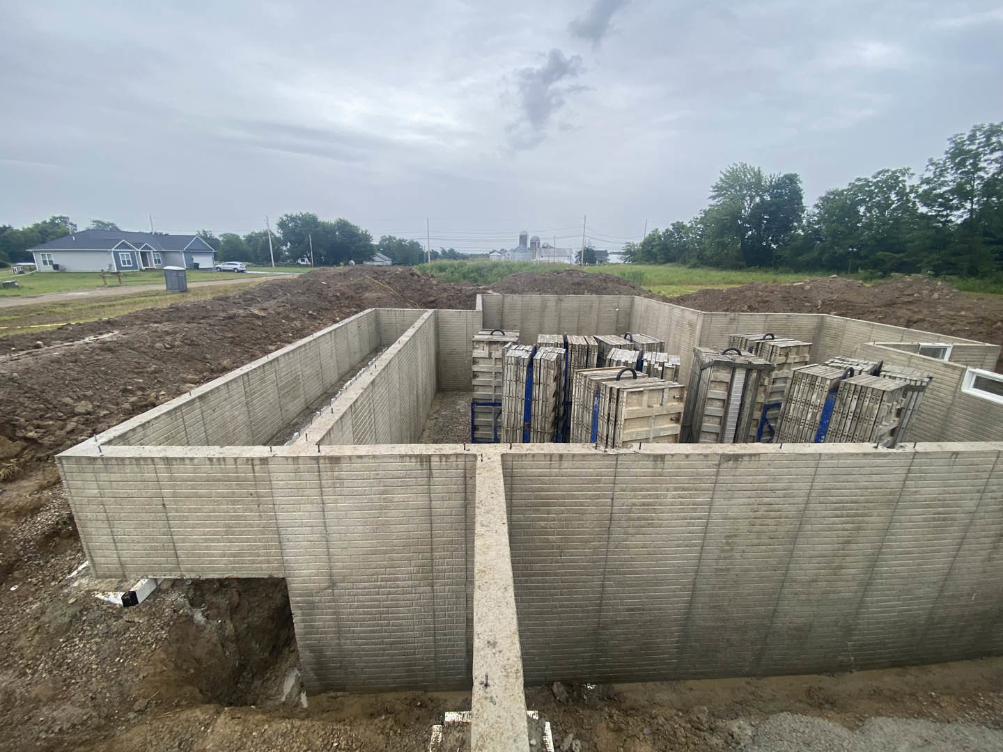 Concrete foundation with exposed soil and metal piles, blue-roofed house in background, cloudy sky, brick wall with white rectangular object, ladder leaning against wall.
