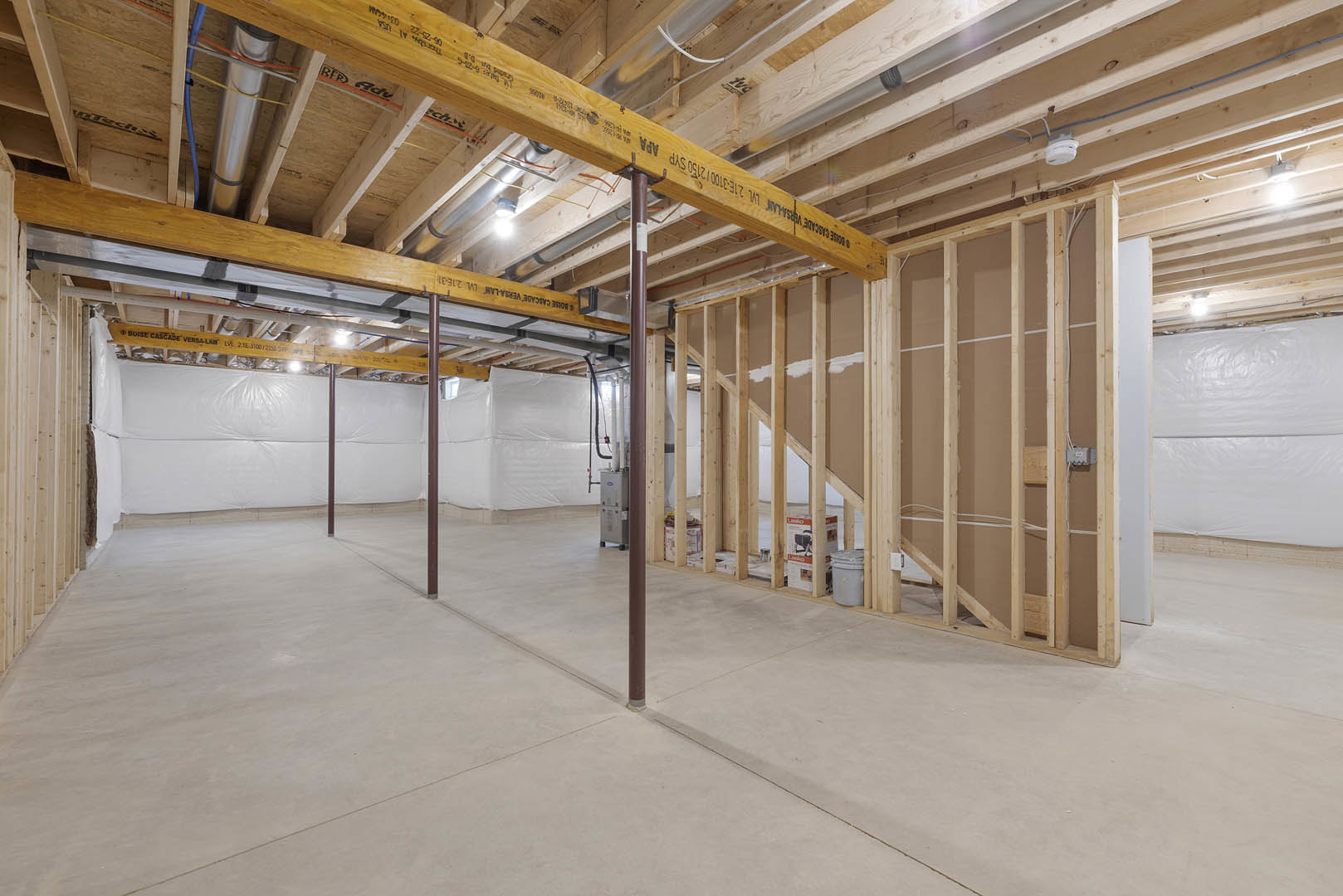 Exposed wooden beam with black markings, concrete floor with metal support poles, white plastic sheet on wooden shelving, visible steel beams, machine with pipes and wires along