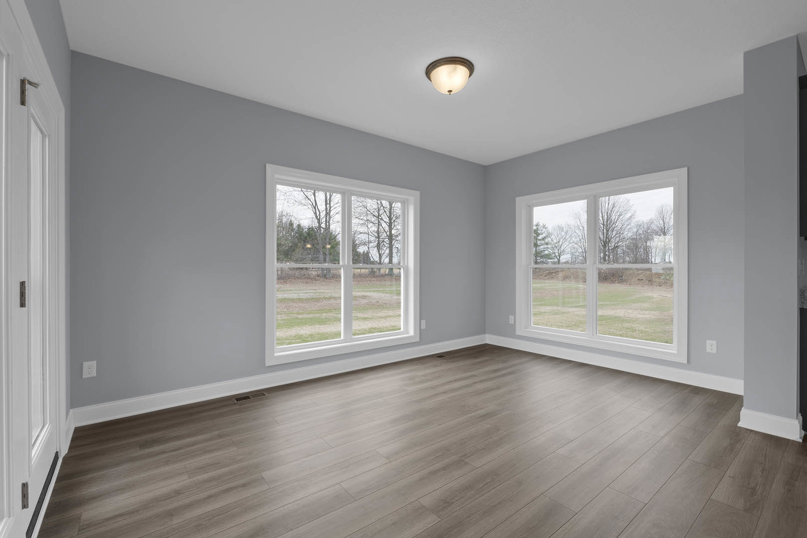Spacious room with hardwood flooring, large windows overlooking trees, white walls, ceiling light fixture, and white door with silver handle