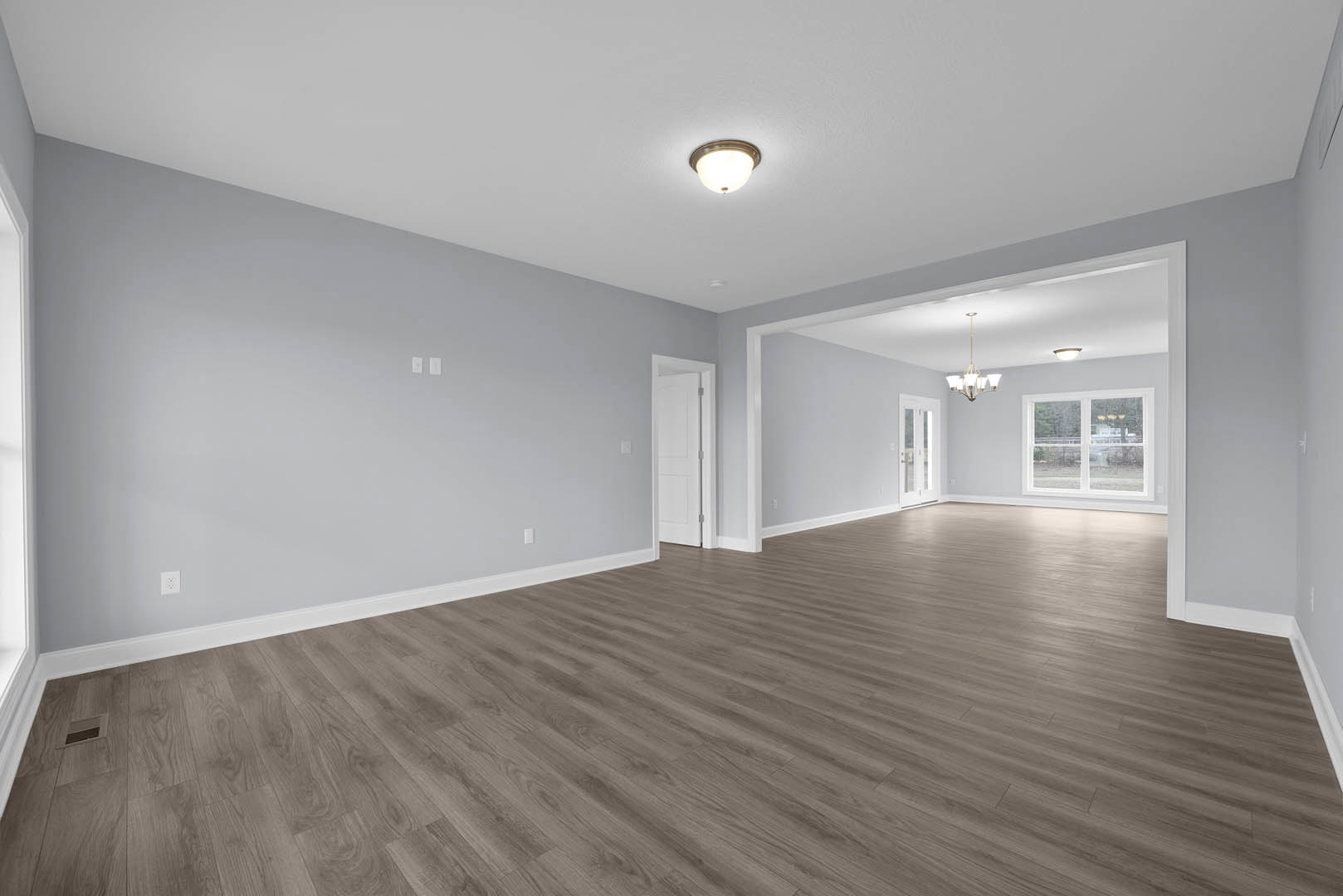 Wood floor room with white walls, ceiling light fixture, window facing neighboring house, and white door with silver handle