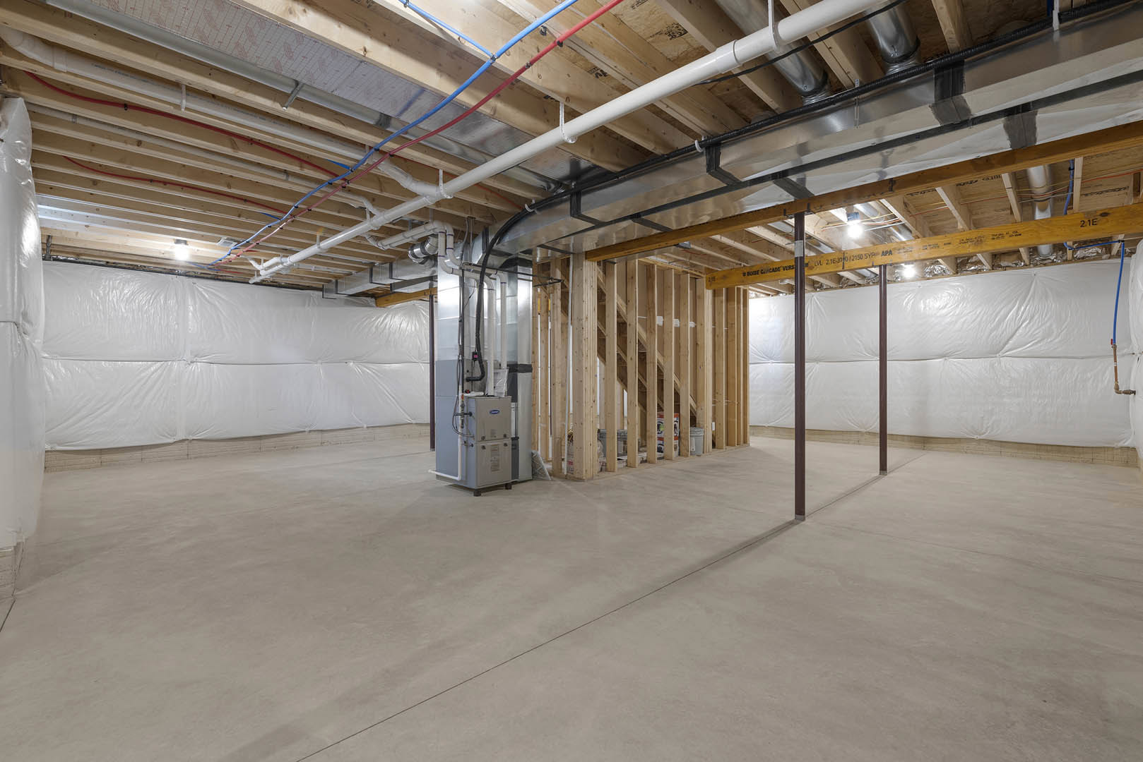Exposed steel beams and pipes run across a high unfinished ceiling above a concrete floor, with a grey utility box, white plastic-covered wall, and building insulation visible in a