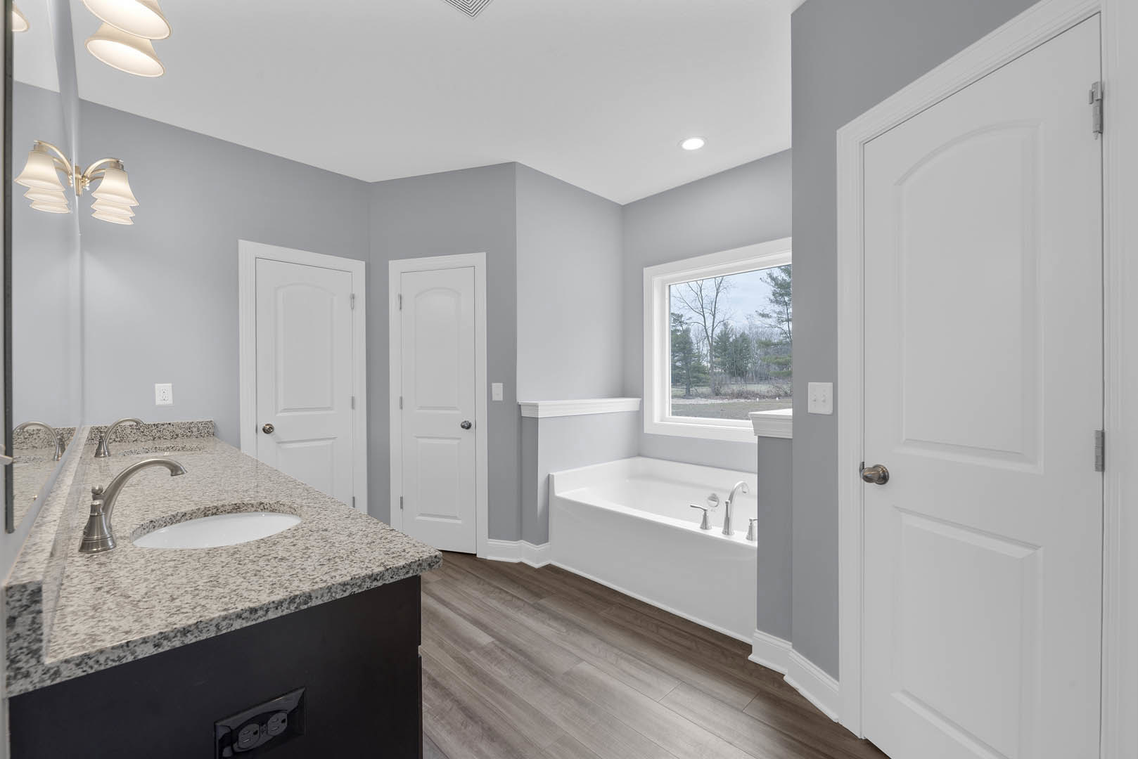 Marble countertop with undermount sink and chrome faucets, freestanding bathtub near window showing trees, white door with silver knob, modern light fixture above, neutral