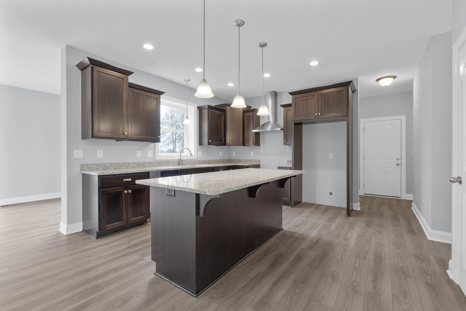 Spacious kitchen featuring a large central island with light stone countertop, wood flooring, white cabinetry with silver hardware, stainless steel faucet over undermount sink, and