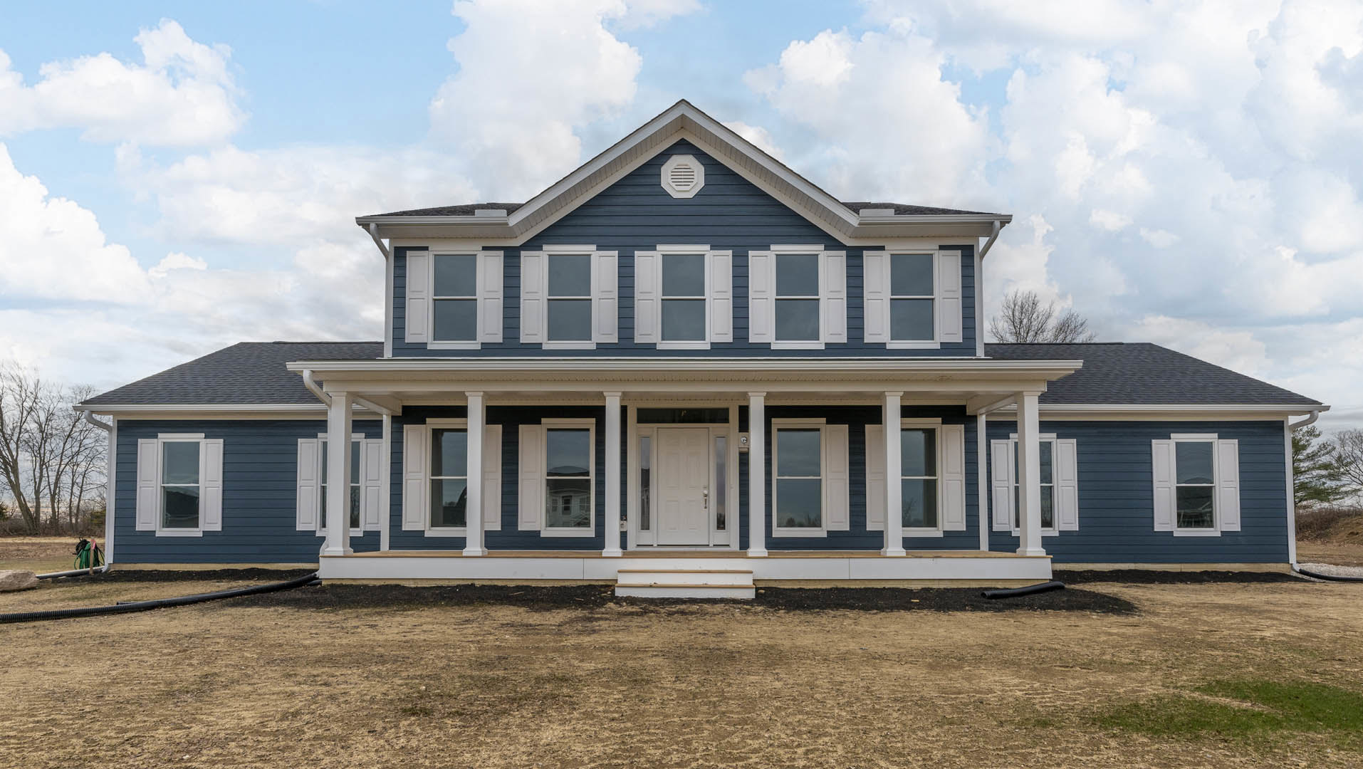 Large front porch with white columns, blue and white front door, glass-paneled windows, vent above entry, steps leading from dirt ground, leafless tree nearby, light-colored siding